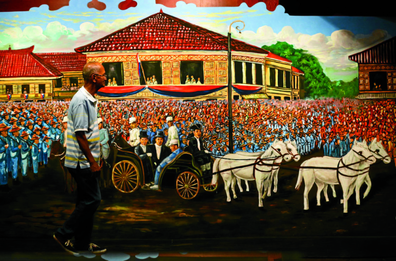 INDEPENDENCE DAY INKAWIT Volunteer tour guide Jose Venerando Vales, 73, shows a mural based on a blackand- white photograph taken in 1899 of Gen. Emilio Aguinaldo holding a fashionable cane while riding a carriage with some companions. The painting at the Aguinaldo Shrine in Kawit, Cavite, depicts Aguinaldo on his way to the inauguration of the First Republic in Malolos Congress.